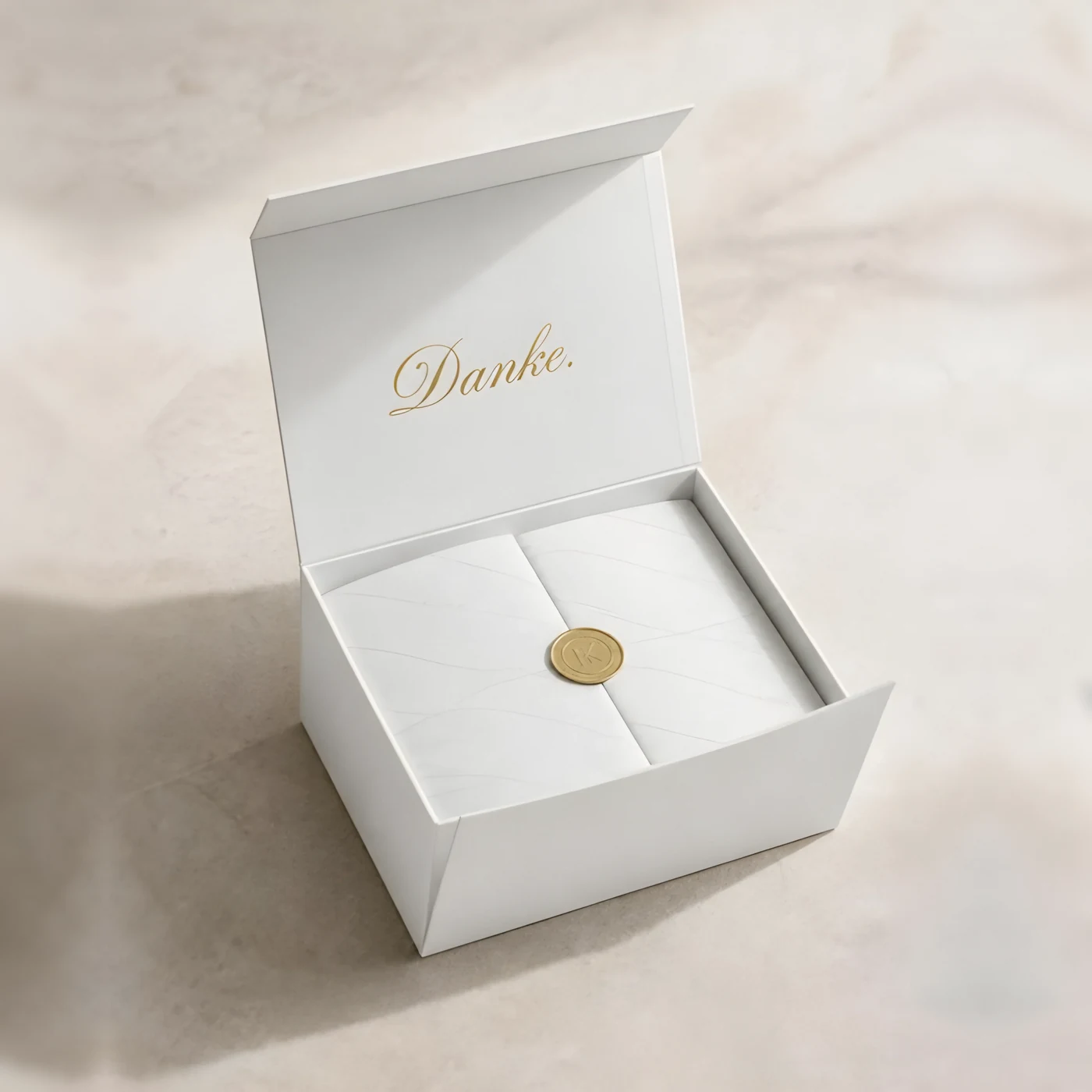 Geöffnete weiße Magnetbox mit „Danke.“ in Goldprägung im Deckel, Inhalt in gemustertem Seidenpapier mit Goldsiegel