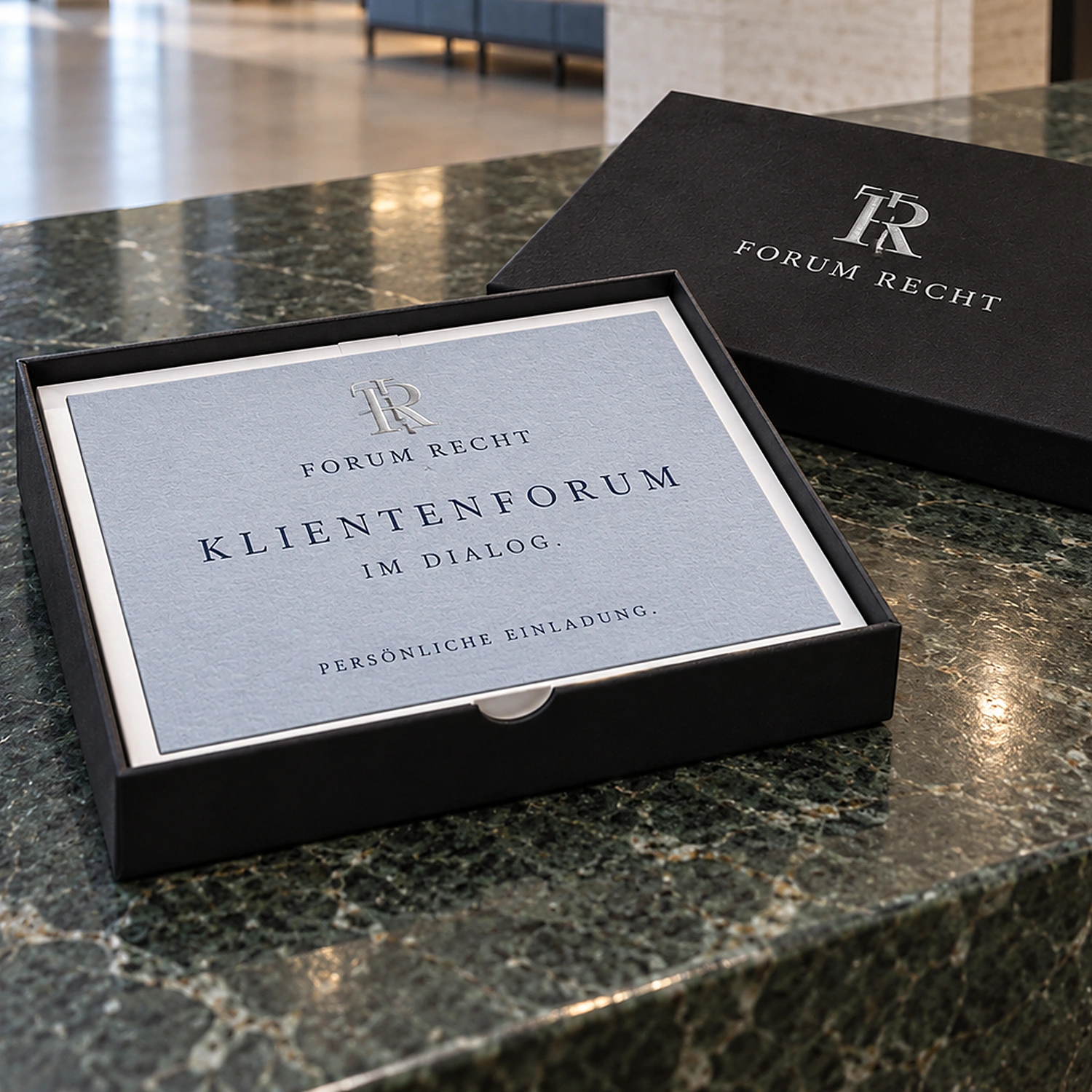 Geöffnete schwarze Stülpdeckelbox mit Einladungskarte auf einem Marmortresen, daneben der Deckel mit Logo in Silber-Heißfolie