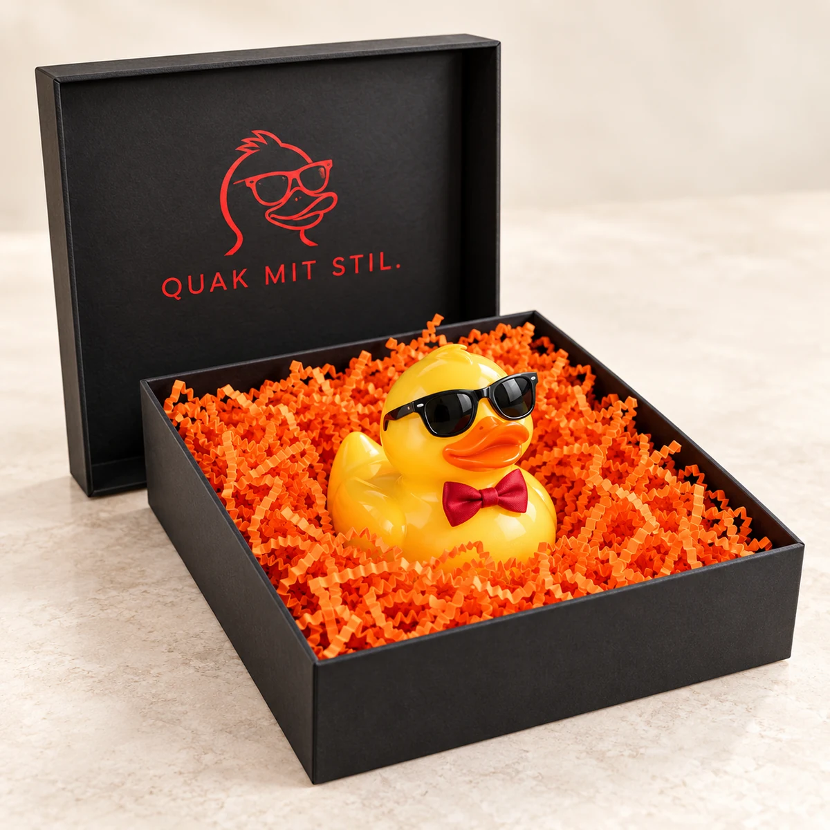 Schwarze Box mit Quietscheente mit Sonnenbrille in orangem Knüllpapier, im Deckel „Quak mit Stil.“ in Rot